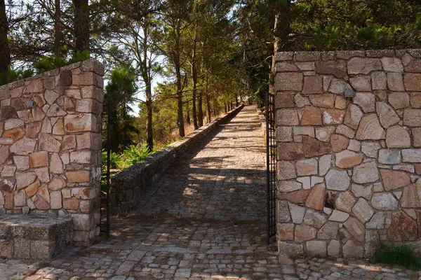 Girişinde kırsal taştan bir kapı bulunan uzun ağaçlarla çevrili sakin bir yol, gölgeli, ağaçlarla kaplı bir koridor boyunca uzanır.