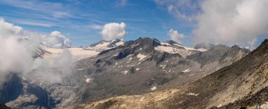 Buzullarla, kayalık arazilerle ve yüksek irtifadaki manzaraların çarpıcı güzelliğini yansıtan bulutlarla dolu görkemli bir dağ sırasının çarpıcı panoramik görüntüsü.