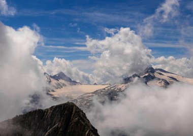 Karla kaplı dağ zirvelerinin bulutlardan oluşan bir battaniyeyi delip geçmesi büyüleyici bir manzara. Parlak mavi gökyüzü, Alp manzarasının ihtişamını vurgulayan sisli atmosferle çelişiyor.