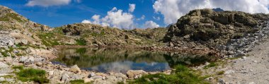 Clear mountain lake with reflections of rocky hills in a high altitude alpine environment, surrounded by pristine nature and panoramic views under a bright blue sky clipart