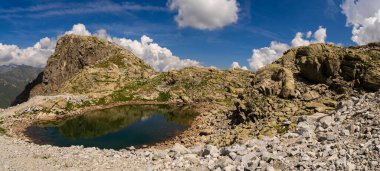 High altitude small alpine lake with clear reflections of rocky cliffs and rugged terrain, surrounded by pristine nature and panoramic mountain views under a bright blue sky clipart