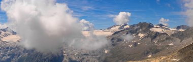 Wide panoramic shot of a mountain range with snow-capped peaks, rocky terrain, and scattered clouds. The clear blue sky enhances the expansive and majestic landscape clipart