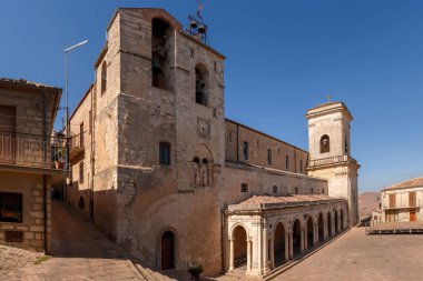 Petralia Soprana, Sicilya 'daki Chiesa dei Santi Apostoli Pietro e Paolo' nun panoramik görüntüsü, taş mimari, saat kulesi ve açık mavi gökyüzünün altındaki köy meydanında kemerli sütunlar.