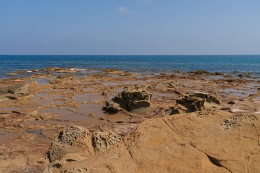 Kayalık bir Akdeniz kıyı şeridinin detaylı bir görüntüsü. Özgün erozyon desenleri ve sığ gel-git havuzları var. Geniş mavi deniz ve açık gökyüzü arka planda.