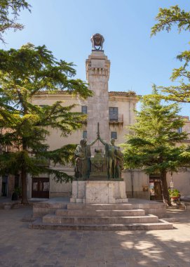 Petralia Soprana, Italy. Monument with statues and a tall stone pillar in the center of a town square, surrounded by trees. The scene is illuminated by bright sunlight under a clear blue sky clipart