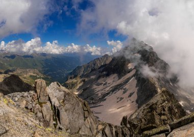 Yüksek tepeler, engebeli kayalar ve sisli bulutlardan oluşan alp manzarasının nefes kesici panoramik görüntüsü. Geniş manzara, canlı mavi bir gökyüzünün altında yemyeşil vadiler boyunca uzanır.