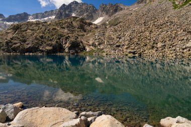 Kristal berrak su ile sakin dağ gölü engebeli kayalık uçurumları ve el değmemiş yaban hayatıyla çevrili sakin dağlık bir çevrede karla kaplı tepeleri yansıtıyor.