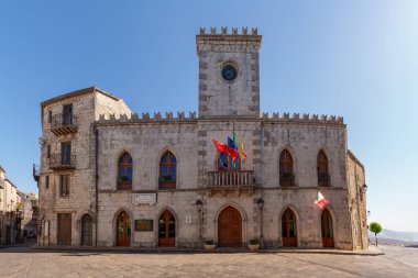 Petralia Soprana, Italy - July, 24, 2024: Grand stone government building with gothic arched windows, central clock tower, colorful flags waving in breeze. The building overlooks a wide town square clipart