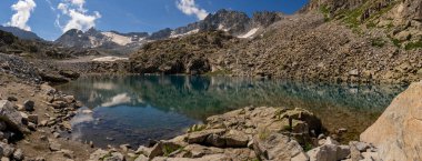 Kayalık araziyle çevrili alp gölünün panoramik manzarası ve mavi gökyüzünün altındaki karlı tepeleri yansıtıyor. Su engebeli dağları yansıtır, yüksek rakımlı manzaranın sakin güzelliğini yakalar.