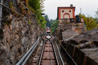 Dik dağ yamacındaki füniküler tren yolu manzarası, arka planda kırsal taş duvarlar ve tuğla bina var.