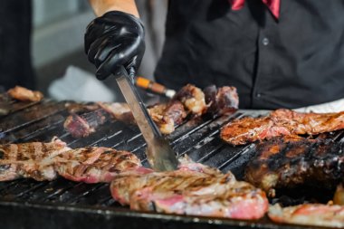 Şef kırmızı bandana takıyor, barbeküde açık ateşte biftek pişiriyor, eti dikkatlice pişiriyor.