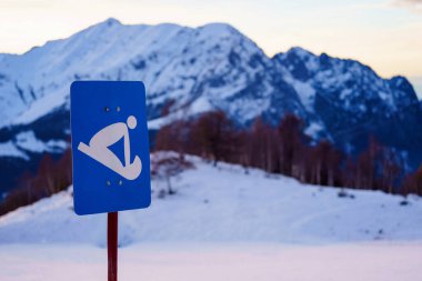 Blue sign indicating sledding, with No Ski No Snowboard written below, stands in front of snow-covered mountain range clipart