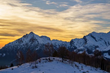 Karla kaplı dağ zirveleri, dağılmış ağaçlar ve dingin gökyüzü ile altın günbatımına karşı dimdik ayakta duruyor. Alp manzarasının dramatik güzelliğini artırıyor, Piani di Bobbio, İtalya