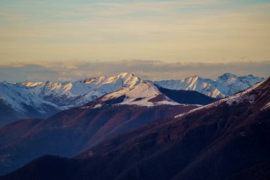 Snow-dusted mountain peaks are illuminated by warm light of sunset near Piani di Bobbio in Italy, creating striking contrast between rugged terrain and the golden sky clipart