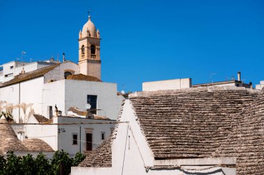 İtalya 'nın Alberobello kentindeki tarihi, beyaz boyalı binalar ve geleneksel taş çatıların çan kulesiyle manzarası. Mimari bu UNESCO Dünya Mirası sahasının eşsiz cazibesini yansıtıyor