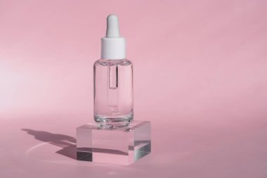 Şeffaf, beyaz kapaklı cam damlalar berrak akrilik bloğun üzerinde duruyor. Yumuşak pembe arka plan, temiz ve minimalist güzellik ürünleri tasarımı.