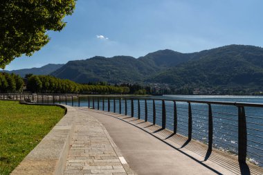 Metal korkuluklu kıvrımlı göl yolu kıyı şeridini takip ederek dağların manzaralı manzarasını ve Lecco Gölü yakınlarındaki sakin suyu sunuyor, Como Gölü. Yol ağaçlarla kaplı, huzurlu bir atmosfer yaratıyor.