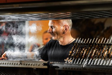 Milan, İtalya - 31.08.2022: Kırmızı bandana takan şef, barbeküde açık ateşte biftek pişiriyor, eti dikkatlice pişiriyor.