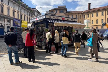 Kartpostallı ve dergili Belmond büfesi. Kalabalık güneşli gökyüzünün altındaki gazete bayiinde geziniyor. Şehir hayatı.