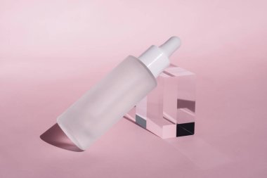 Şeffaf, beyaz kapaklı cam damlalar berrak akrilik bloğun üzerinde duruyor. Yumuşak pembe arka plan, temiz ve minimalist güzellik ürünleri tasarımı.