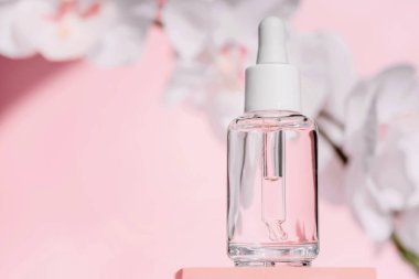 Şeffaf, beyaz kapaklı cam damlalar berrak akrilik bloğun üzerinde duruyor. Yumuşak pembe arka plan, temiz ve minimalist güzellik ürünleri tasarımı.