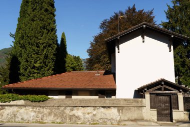 Kırmızı kiremit çatılı beyaz ev taştan duvarın arkasında uzun selvi ağaçlarıyla çevrili. Binanın ahşap kapısı var ve öğleden sonra güneşiyle aydınlanıyor. Ağaçlar ve açık gökyüzü var.