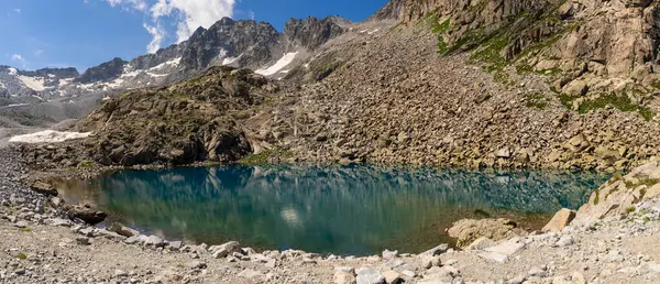 Durgun dağ gölü kristal berrak su ile kayalık kayalıkları yansıtıyor ve el değmemiş dağlık bir çevrede kar kaplı zirveler, dokunulmamış doğayı ve engebeli güzelliği sergiliyor.