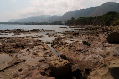 Gelgit havuzları olan kayalık bir kıyı manzarası, uzak tepeler ve bulutlu bir gökyüzü üzerine kurulmuş, deniz kenarının doğal güzelliğini yansıtıyor.