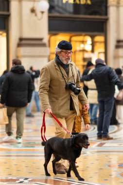 Milan, İtalya - 18.12.2022: Bej ceketli bir adam Victor Emmanuel 'in galerisinde siyah köpeğini gezdirir. Zarif ortam ve çevreleyen kalabalık rahat ama sofistike şehir atmosferi yaratıyor.