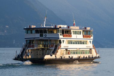 Varenna, İtalya - 01.08.2023: Yolcu feribotu alacakaranlıkta Como gölünün sakin sularında, yüksek dağlarla çevrili, göl manzarasında sakin bir an
