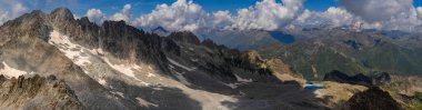 Geniş bir panoramik çekim parçalı bulutlu bir gökyüzünün altında engebeli dağ sıralarını kar ve kayalık arazi parçalarıyla yakalıyor..