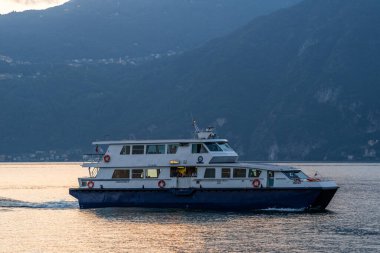 Varenna, İtalya - 01.08.2023: Feribot gün batımında Como Gölü 'nün sakin sularında süzülür ve arka planda dağlık bir manzara vardır. Huzurlu akşam manzarası dingin gölü vurguluyor