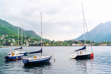 Lecco, İtalya - 01.05.2023: Bereketli yeşil tepeler ve arka planda manzaralı bir kasaba, İtalya 'nın Como Gölü yakınlarındaki Lecco yakınlarında huzurlu bir deniz kenti olan sakin göle demirlemiş yelkenliler
