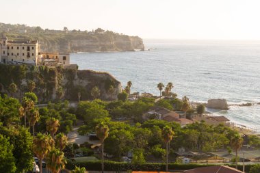 Tropea, İtalya - 27.07.2023: Kıyı şeridi manzarası, yemyeşil ağaçlar, palmiye ağaçları, uçurumlardaki tarihi yapılar ve öğleden sonra arkaplanda parıldayan okyanus manzarası