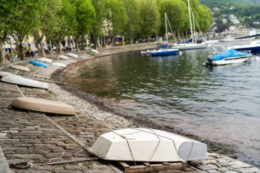Lecco, İtalya - 01.05.2023: Ters dönmüş kayıklar arka planda sakin göle demirlemiş yelkenlilerle kıyıda dinleniyor. İtalya 'daki Como Gölü' nde Lecco yakınlarında huzurlu bir açık hava sahnesi.