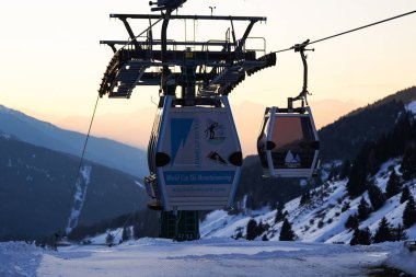 Passo Tonale, İtalya - 28.01.2023: Karlı dağ manzarası üzerinde hareket eden Gondol, günbatımının yumuşak ışığıyla aydınlanıyor ve arka planda görünen dağlık tepeler, alp kayak merkezi