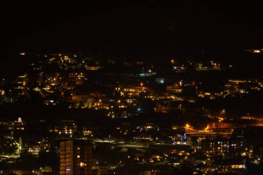 Gece yarısı aydınlık binaları ve caddeleri olan bir tepe kenti manzarası sıcak ve canlı bir atmosfer yaratıyor. Kentsel, gece ve seyahat temaları için ideal