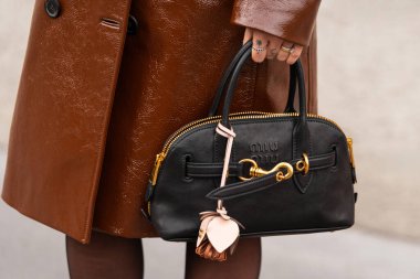 Paris, Fransa - 11 Mart 2025: Siyah Miu Miu çanta altın ayrıntılar ve cazibe ile. Kahverengi parlak patent trençkotlu. Sokak tarzı kıyafet ayrıntıları.