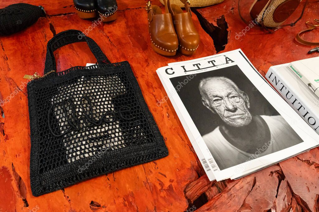Milan, Italy - April, 08, 2025: Chloe black mesh crochet tote bag, clogs, and fashion books on textured red table. Boutique retail detail, Milan showroom.