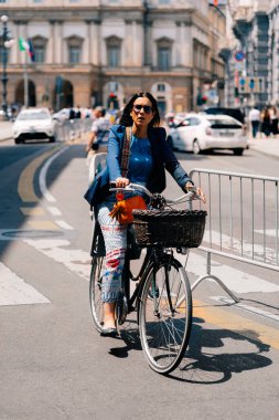 Milan, İtalya - 25 Mayıs 2024: Mavi ceketli ve renkli pantolonlu şehir caddesinde bisiklet süren kendinden emin bir kadın. Sürdürülebilir seyahat kavramı, Milan tarzı