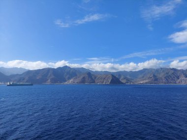 A breathtaking view of an expansive blue ocean meeting rugged mountains under a clear sky. A ship is seen sailing on the horizon, highlighting the beauty of nature and exploration. clipart