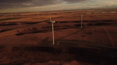 Aerial windmills producing sustainable energy on rolling hills at sunset with distant Canadian Rocky Mountains.