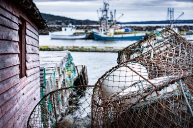 Lobster traps and crab traps stacked on a dock next to a tackle shed with fishing trawlers at background at Whiteway Newfoundland Canada. clipart