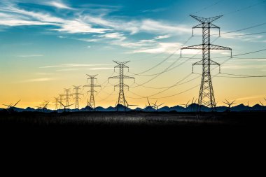 Uzak dağlara ve değirmenlere tepeden bakan çelik kafes şanzıman kuleleri ve elektrik hatlarından oluşan gün batımı silueti.