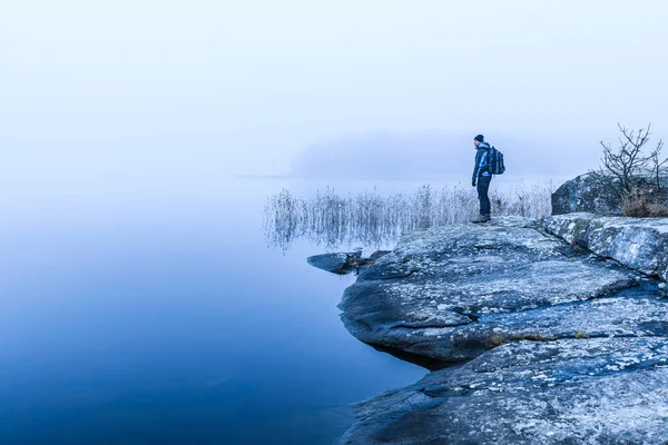 A lone hiker admires the beauty of nature reflected in a tranquil lake, surrounded by misty skies in Sweden.