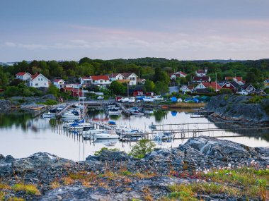 İsveç 'te sakin bir liman sahnesi, nehir kıyısı boyunca huzur içinde dinlenen tekneler, yemyeşil bitki ve binaların olduğu bir zemin..