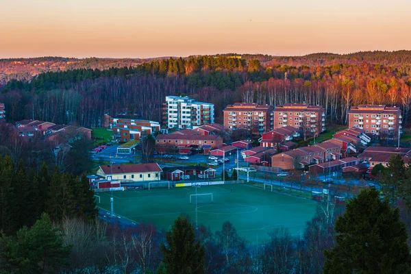 At dusk, the orange sky casts a dreamy glow over residential area; buildings, trees and a football field peacefully coexist in this scenic cityscape.