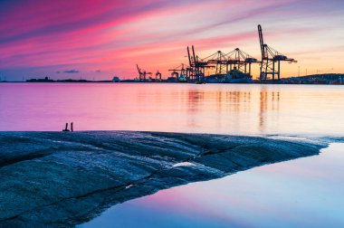 Günbatımında ticari bir liman: kargo konteynerleri, vinçler ve bir denizcilik gemisi.