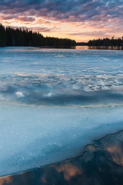 Huzurlu bir sahne alacakaranlıkta kısmen donmuş bir gölün güzelliğini yakalar. Gün batımının ince renkleri etrafındaki ormanın ortasında buzu ve suyu yansıtır..