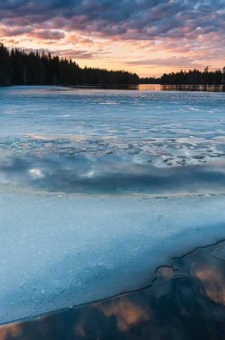 Huzurlu bir sahne alacakaranlıkta kısmen donmuş bir gölün güzelliğini yakalar. Gün batımının ince renkleri etrafındaki ormanın ortasında buzu ve suyu yansıtır..
